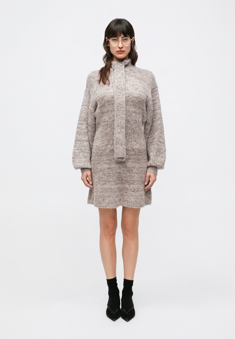 Pletené šaty v jemnej béžovej farbe s nákrčníkom a dlhými rukávmi. Má priamy strih, jemnú textúru a viazanie na krku.