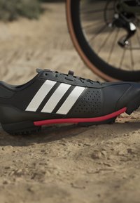 Černá sportovní bota se třemi bílými proužky, texturovaný síťovaný svršek, růžové detaily na podešvi a malé hroty pro grip na písčitém terénu.