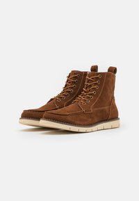 Botas de tobillo de ante marrón con diseño de cordones y ojales de metal. Presentan una suela de goma crema y costuras visibles a lo largo del borde.