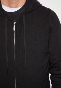 Czarny bluz z kapturem na zamek, wykonany z teksturowanej tkaniny, z suwakiem z przodu, sznurkami do regulacji oraz warstwowym wyglądem z białym T-shirtem pod spodem.
