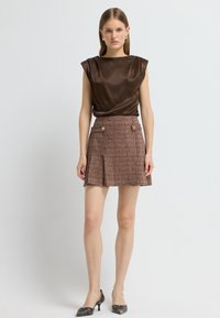 Haut en satin brun avec épaules structurées, associé à une mini-jupe en tweed dans des tons bruns, ornée de deux boutons dorés et de plis. Talons noirs.