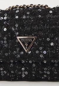Borsa nera con texture di paillettes, caratterizzata da un logo triangolare Guess in argento e una tracolla in oro. Design piatto e rettangolare.