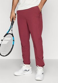 Sarkani sporta bikses, izgatavotas no viegla materiāla, ar sānu kabatām, šauru piegriezumu un baltu akcentu šuvēm gar piedurknēm.