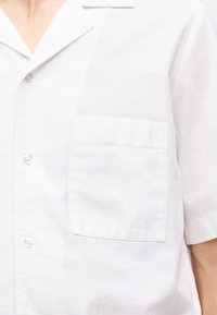 Chemise blanche à manches courtes avec une poche poitrine et des boutons, portée par une personne avec le cou et le bras visibles.