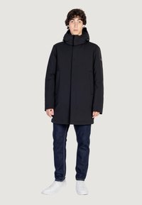 Cappotto imbottito nero con colletto alto, chiusura frontale con zip e tasche laterali. Indossato sopra jeans scuri e sneakers bianche. Design semplice e moderno.