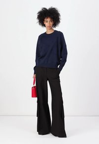 Mørkeblå genser med teksturert mønster, kombinert med høytlivede svarte vide bukser. Modellen bærer en liten rød skulderveske.