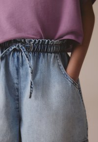 Lyseblå denimshorts med elastik i taljen og bindedetalje, kombineret med en løstsiddende lilla skjorte. Synlig lomme på højre side.