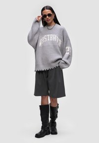 Szary, oversize wełniany sweter z napisem "WRSTBHVR" i postrzępionym dołem, w połączeniu z ciemnoszarymi szortami oraz czarnymi, wysokimi butami na kolana z klamrami.