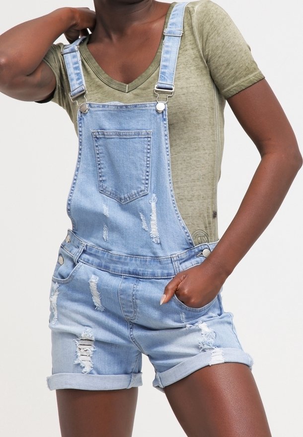 Hellblaue Latzhose mit distressed Details, getragen über einem grünen V-Ausschnitt-T-Shirt. Mit verstellbaren Trägern und hochgekrempeltem Saum.