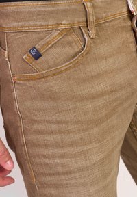 Khaki-kleurige jeans met een gladde textuur, voorzien van oranje stiksel en een klein merkteken op de voorkant van de zak.