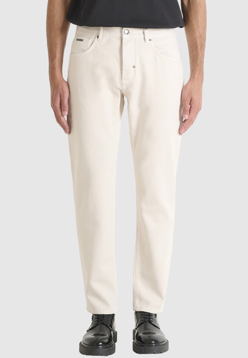 Hombre vestido con jeans rectos color blanco roto y botas de cuero negro con cordones, de pie contra un fondo claro y liso.
