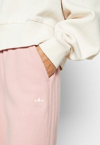 Pantalones de chándal de color rosa claro con una textura suave, que presentan tres rayas blancas a lo largo del costado y un logo bordado en el muslo.