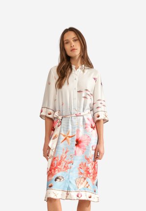 Nainen pukeutuneena polvipituiseen, napitettuun mekkoon, jossa on merieläinkuvioita, kuten tähdenmuotoisia meritähtiä, koralleja, simpukoita ja kukkia, poseeraa yksiväristä taustaa vasten.