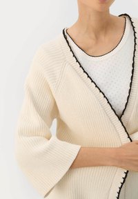 Krémový pletený cardigan s černými vroubkovanými okraji, volnými rukávy a texturovanou tkaninou; přeložený přes bílou perforovanou blůzu.
