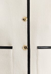 Biała kurtka z mieszanki wełny z czarnymi skórzanymi akcentami. Posiada złote guziki z logo oraz dwie przednie kieszenie obramowane na czarno. Gładka tekstura.