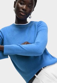 Modrý pletený svetr s kulatým výstřihem a ribovanými manžetami, který má hladký povrch. Nesený na bílých kalhotách s čistými liniemi.