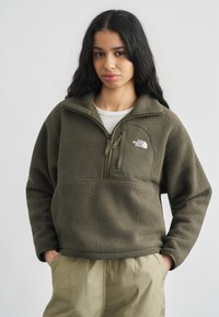 Γυναίκα με μακριά σκούρα μαλλιά που φοράει πράσινη φλις μπλούζα με φερμουάρ στην τσέπη στο στήθος και μπεζ παντελόνι, στέκεται μπροστά σε απλό φόντο.