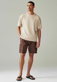 Beige T-Shirt mit kurzen Ärmeln, braune Cargo-Shorts mit Seitentaschen und schwarze Flip-Flops. Der Stoff wirkt weich und hat eine lässige Passform.