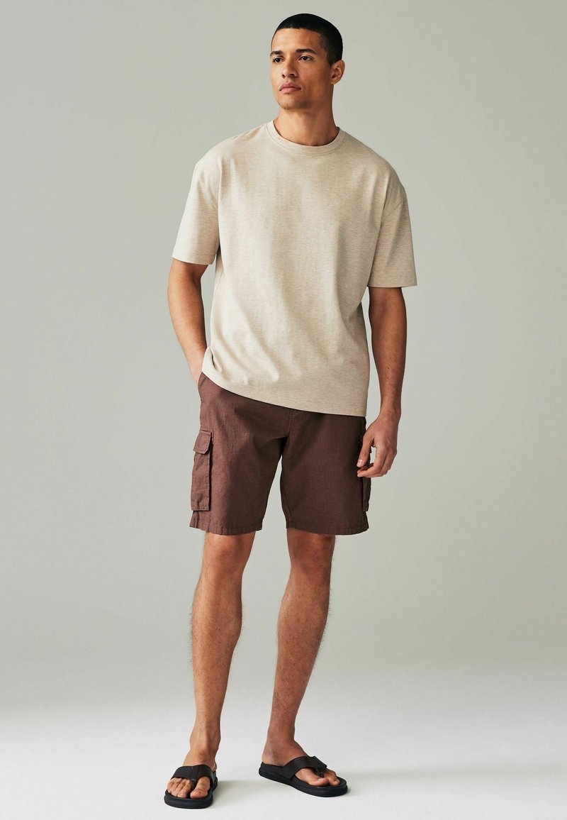 Beige T-Shirt mit kurzen Ärmeln, braune Cargo-Shorts mit Seitentaschen und schwarze Flip-Flops. Der Stoff wirkt weich und hat eine lässige Passform.