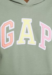 Sudadera con capucha verde con letras bordadas multicolores que forman "GAP" en rosa, amarillo y naranja. Textura suave y detalle de bolsillo tipo canguro.