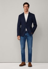 Marineblazer über einem weißen Hemd, kombiniert mit hellblauen Jeans und braunen Schuhen. Der Blazer hat einen einzigen Knopf und Fronttaschen.