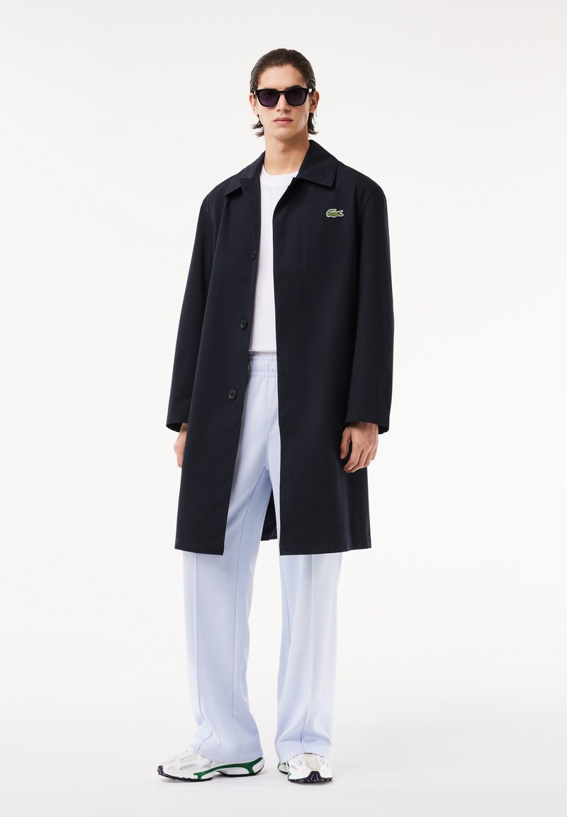 Largo abrigo negro con botones, llevado sobre una camisa blanca y pantalones azul claro. Presenta un logo verde bordado en el pecho. Gafas de sol negras.