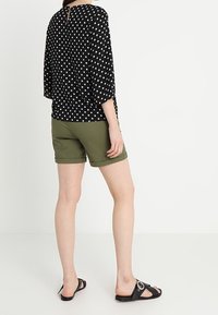 Kvinna iförd en svart och vit prickig topp, olivgröna uppvikta shorts och svarta slidesandaler, stående mot en enkel bakgrund.