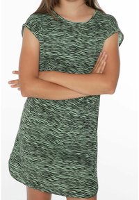 Barn med korslagda armar som bär en kortärmad, zebrarandig grön och svart klänning mot en vit bakgrund.