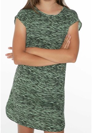 Enfant les bras croisés portant une robe à manches courtes rayée de zèbre verte et noire devant un fond blanc.