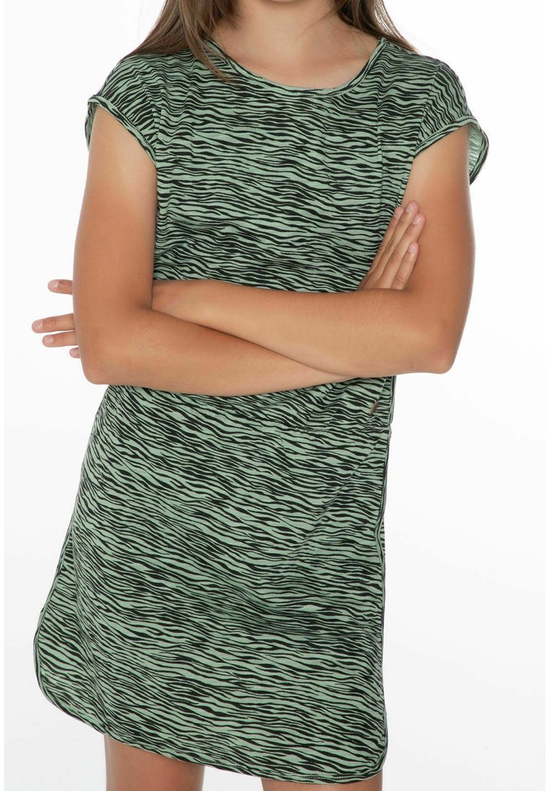 Barn med korslagda armar som bär en kortärmad, zebrarandig grön och svart klänning mot en vit bakgrund.