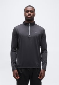 Άνδρας που φοράει μαύρο γκρι μπλουζάκι με μακριά μανίκια και φερμουάρ ως τη μέση, με το λογότυπο "ck" στο στήθος, στέκεται μπροστά σε απλό λευκό φόντο.