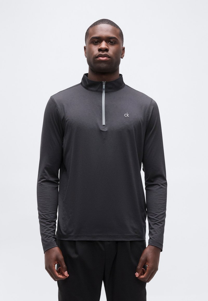 Άνδρας που φοράει μαύρο γκρι μπλουζάκι με μακριά μανίκια και φερμουάρ ως τη μέση, με το λογότυπο "ck" στο στήθος, στέκεται μπροστά σε απλό λευκό φόντο.