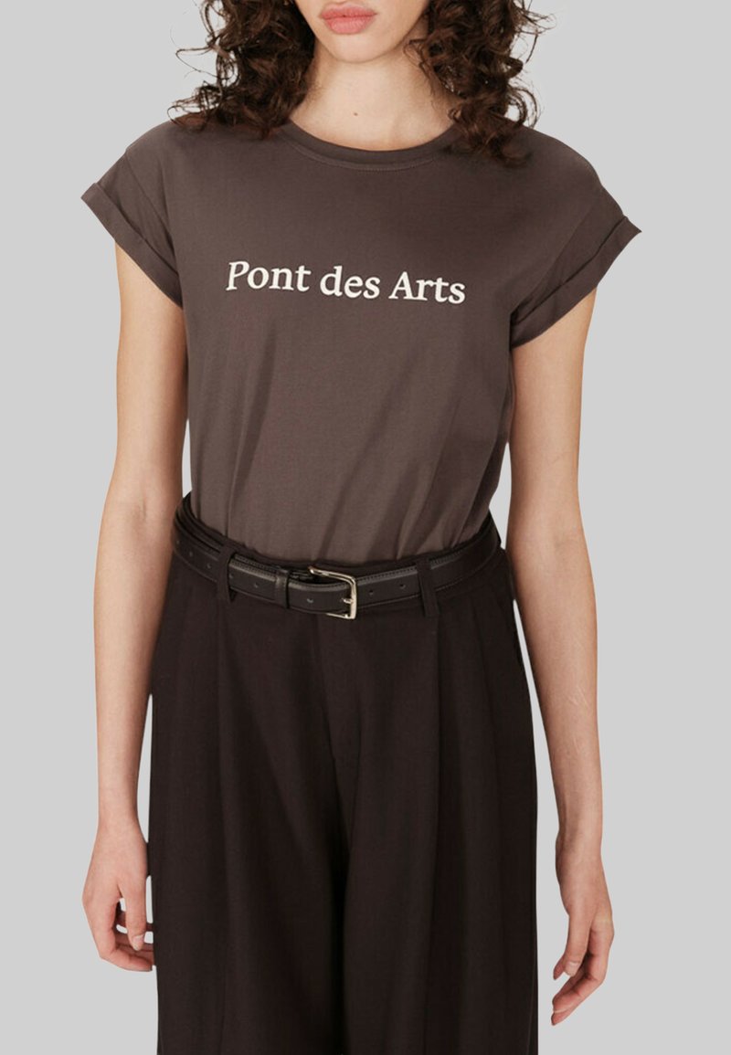 T-shirt en coton marron avec des manches retroussées et le texte blanc "Pont des Arts." Porté avec un pantalon noir taille haute et une ceinture noire.