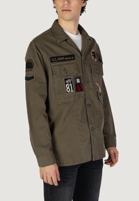 Olijfgroene militaire stijl shirtjacket, met meerdere geborduurde patches, twee borstzakken, een knoopsluiting en een klassieke kraag.