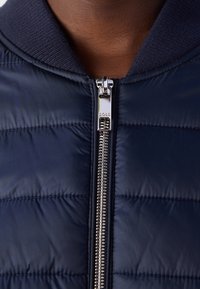 Close-up van een persoon die een marineblauwe doorgestikte jas draagt met een zilveren ritssluiting en een geribde kraag, dichtgeritst tot aan de hals.