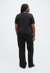 Γυναίκα με μακριά πλεξούδα που φοράει μαύρο κοντομάνικο πουκάμισο, μαύρο παντελόνι με φαρδιά πόδια και καφέ παπούτσια, στέκεται γυρισμένη προς τα πίσω.