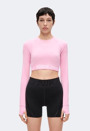 Mujer vistiendo un top corto rosa de manga larga y pantalones cortos negros de cintura alta, de pie mirando hacia adelante con una expresión neutral sobre un fondo liso.