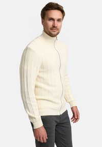 Weißer, gerippter Pullover mit Reißverschluss, hohem Kragen und langen Ärmeln, kombiniert mit dunkelgrauer Hose. Verfügt über eine strukturierte Strickoptik und einen Frontreißverschluss.