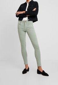 Jeans ajustados en una mezcla de algodón verde claro, que cuentan con cinco bolsillos, pasadores para cinturón y sutiles detalles de costura, combinados con una chaqueta de mezclilla negra.