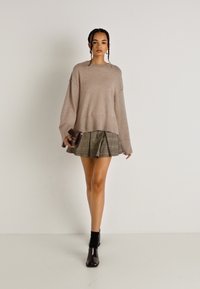 Μπεζ πλεκτό πουλόβερ με άνετη εφαρμογή, συνδυασμένο με πλεχτή μίνι φούστα σε ελαιόχρωμο καρό και μαύρες μπότες μέχρι τον αστράγαλο, κρατώντας μια μικρή σκοτεινή τσάντα.