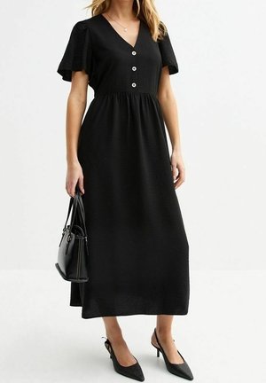 Vrouw draagt een zwarte midijurk met korte vleugelmouwen, drie knopen aan de voorkant, zwarte puntige hakken en houdt een zwarte handtas vast.
