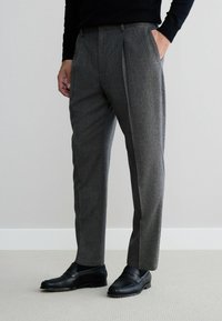 Graue, gefaltete Hose aus strukturiertem Stoff, mit Seitentaschen, kombiniert mit schwarzen Loafers und einem eng anliegenden schwarzen Oberteil.