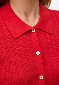 Röd stickad cardigan med en scalloped knappslå, prydd med små vita knappar och en klassisk krage. Texturerad med öppna stygn.