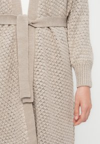 Langer, offen gestrickter Cardigan in hellem Beige mit passendem Bindegürtel. Mit gerippten Bündchen und einem strukturierten Stoffmuster.