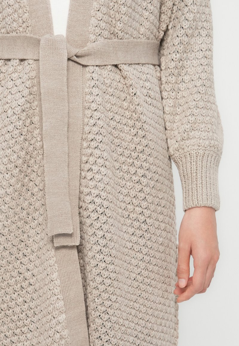 Cardigan long en maille ouverte beige clair avec une ceinture assortie. Présente des poignets côtelés et un motif de tissu texturé.