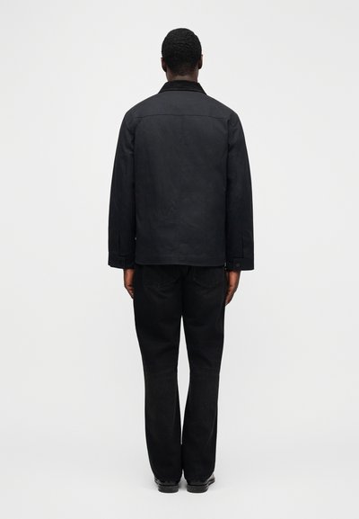 Μαύρο σακάκι από καμβά με γιακά, μανσέτες με κουμπιά και λεπτομέρεια πλάτης, συνδυασμένο με μαύρο τζιν σε φαρδιά γραμμή και μαύρα παπούτσια.