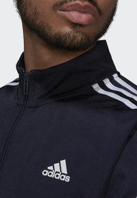 Chaqueta de zip azul marino con diseño de tres rayas blancas en los hombros. Presenta una tela texturizada y un pequeño logo blanco de Adidas en el pecho.