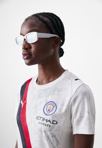 Fehér sportpóló Manchester City logóval, piros és fekete csíkokkal, texturált mintával, és párosítva túlnőtt fehér napszemüveggel.