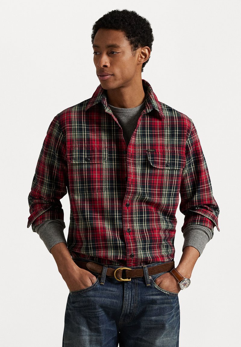 Camisa de franela a cuadros rojos y negros con puños grises, dos bolsillos en el pecho, cierre de botones, combinada con jeans azules y un cinturón marrón.