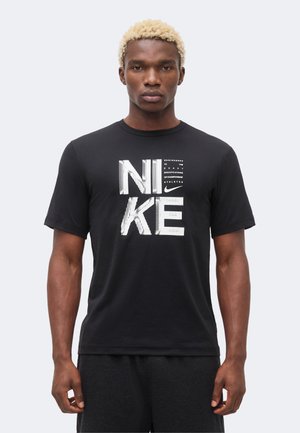 Młody mężczyzna z krótkimi blond włosami, ubrany w czarną koszulkę Nike z białym napisem i logo "NIKE", stojący na jednolitym jasnym tle.